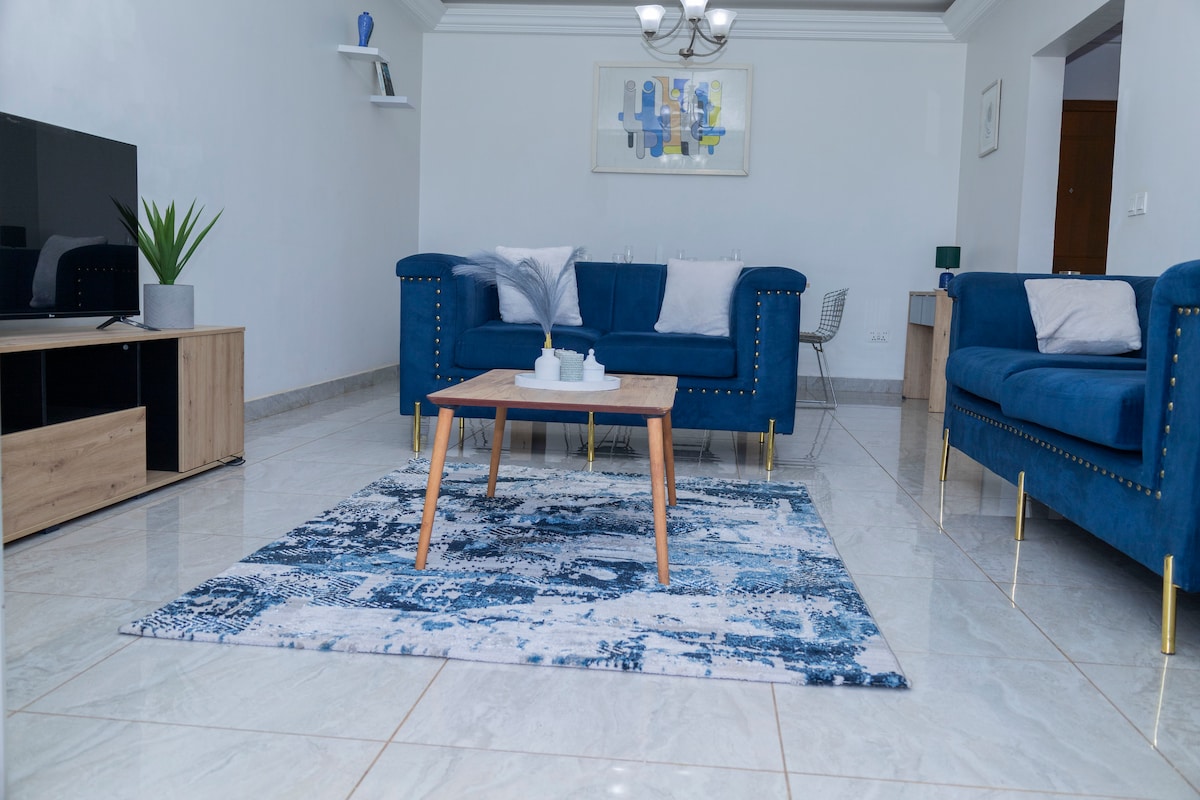 A spacious living area features two blue sofas with decorative cushions arranged around a wooden coffee table. A patterned rug lies on the tiled floor, complemented by a flat-screen TV on an entertainment unit. Natural light fills the room from nearby windows.