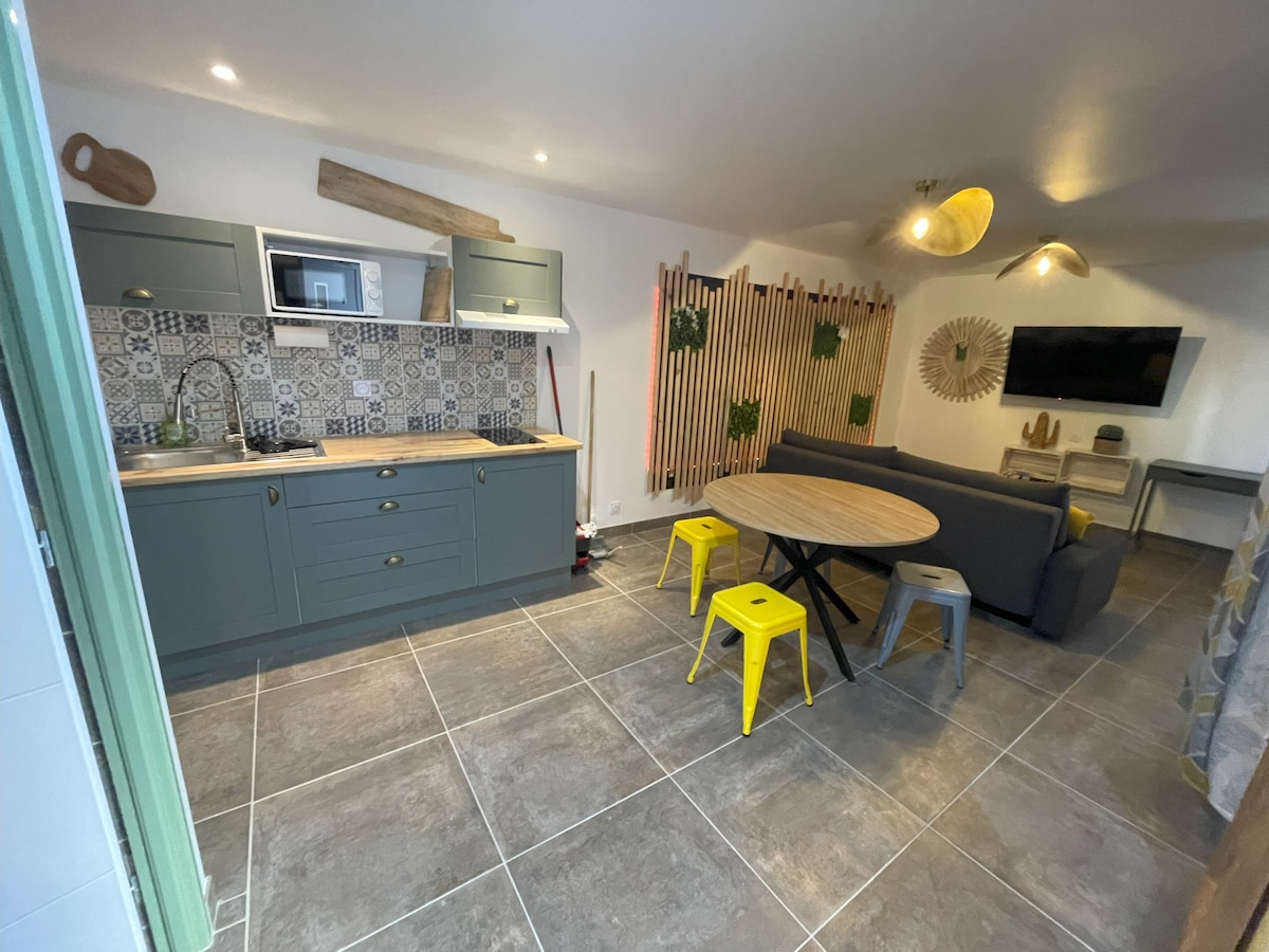 An open-plan living space is revealed, featuring a modern kitchen with grey cabinetry and decorative tiles. A round table with yellow stools contrasts against a backdrop of a grey sofa. Greenery is present along the wall, and a television is mounted within the room.