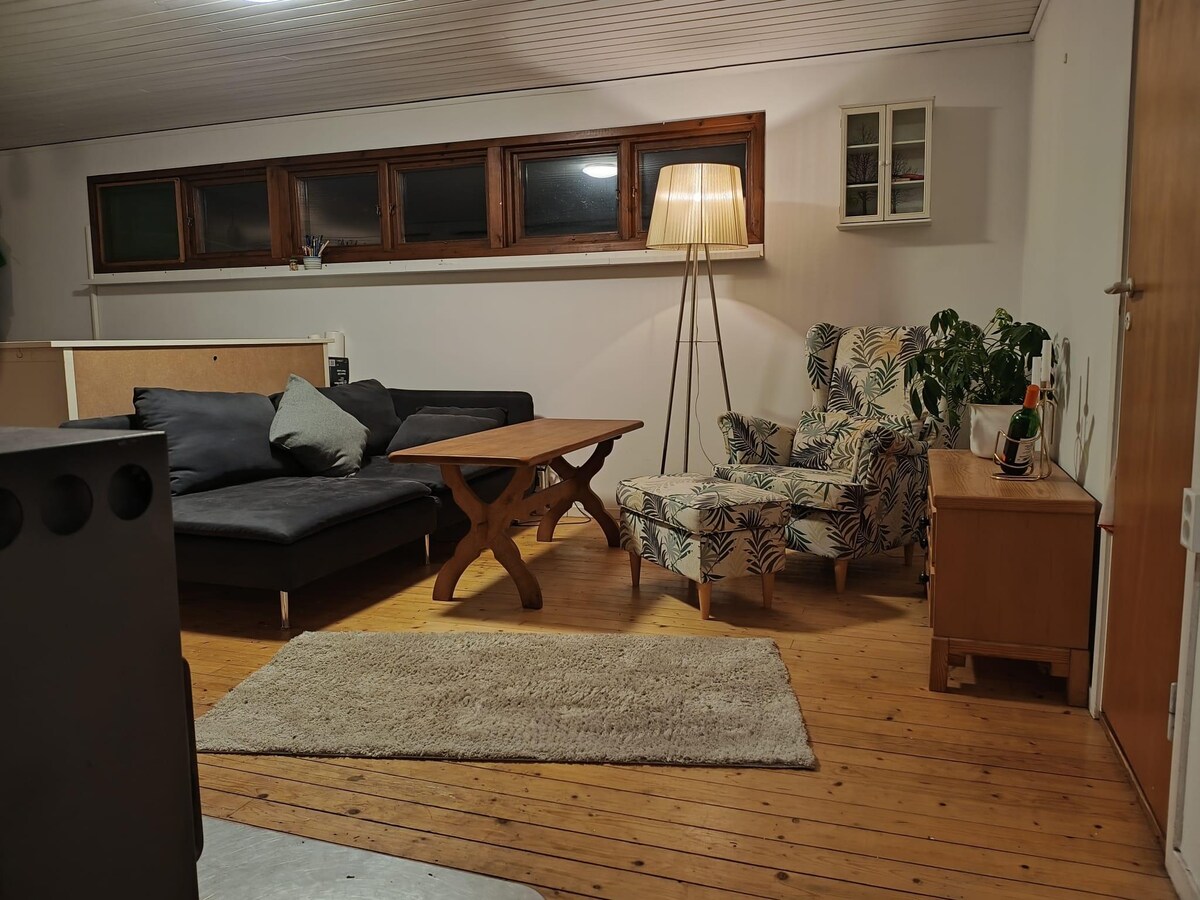 A cozy living area features a dark sectional sofa complemented by a rustic wooden coffee table. A patterned armchair adds a touch of character, while a soft rug anchors the space. Tall windows allow natural light to enhance the warm wooden flooring.
