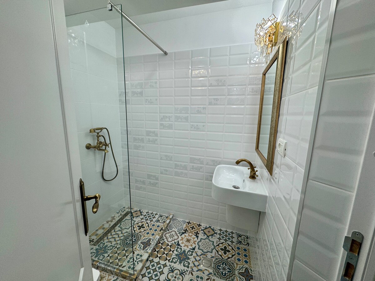 The bathroom features a walk-in shower with a glass enclosure and gold fixtures. A modern sink is positioned beside a decorative mirror. The floor showcases an intricate patterned tile design, contrasting with the clean white wall tiles, enhancing the space's contemporary look.
