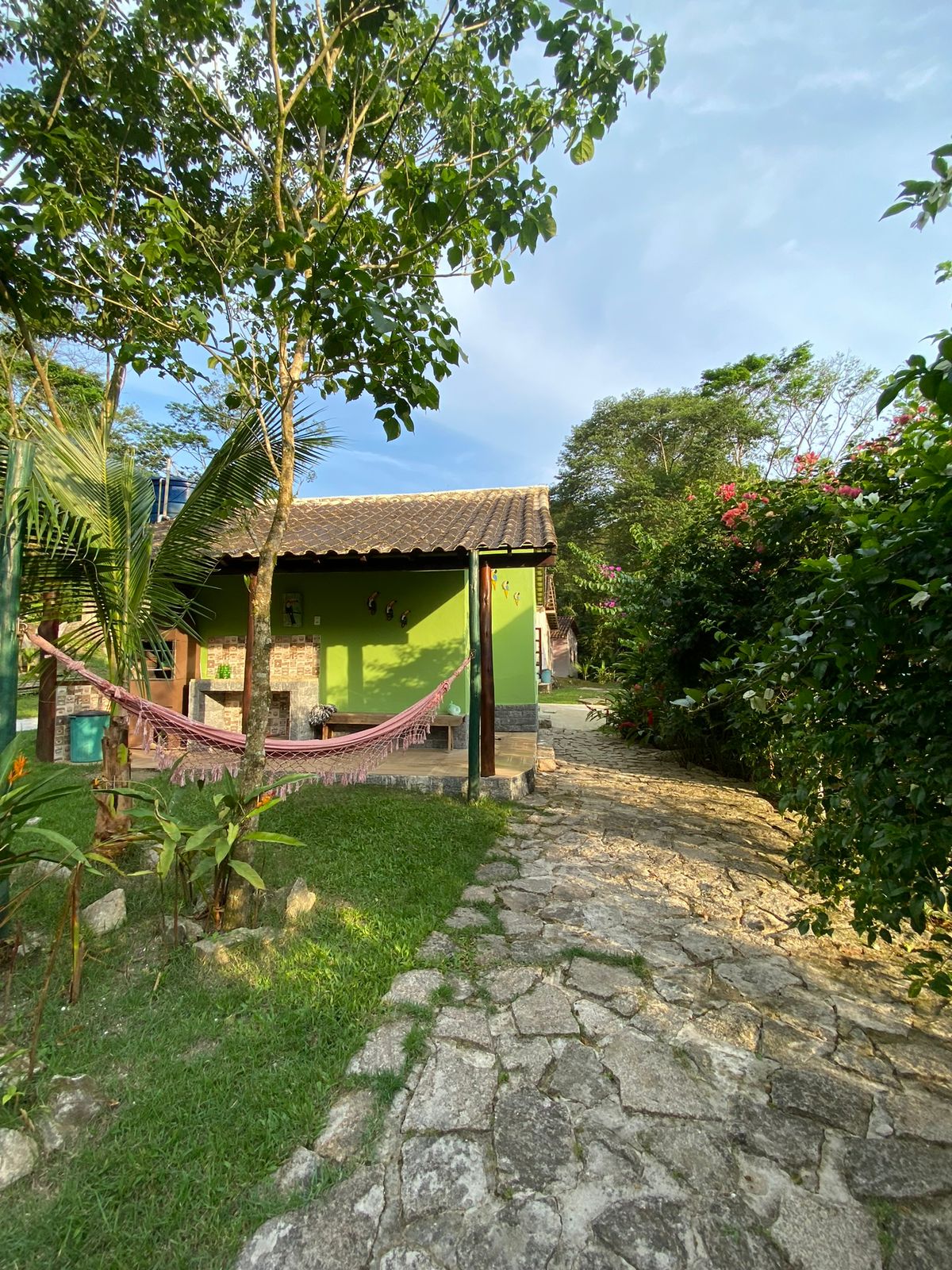 A charming path leads to a green-walled chalet, framed by lush plants and vibrant flowers. A hammock hangs invitingly on the porch, encouraging relaxation. The natural stone pathway is bordered by grassy areas, offering a serene atmosphere embraced by the surrounding greenery.