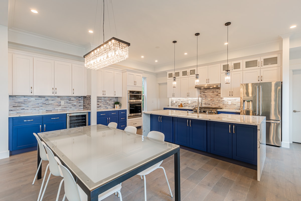 The kitchen features a modern design with white cabinetry and blue accents. A large island counter is surrounded by several chairs. Under-cabinet lights illuminate the workspace, and a stylish chandelier hangs above the dining table, enhancing the contemporary atmosphere.