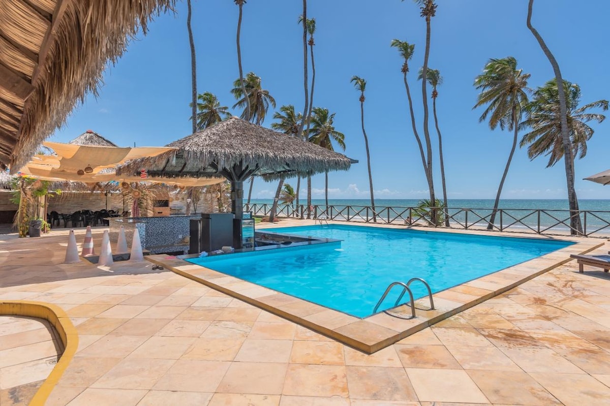 An inviting outdoor area features a clear blue swimming pool surrounded by tropical palm trees. Thatched roofing provides shade for a nearby seating area. The ocean is visible in the background, enhancing the beachside setting.