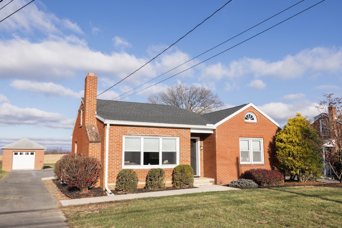 A renovated brick home is shown under a clear blue sky, featuring large windows and a distinctive front arch. The manicured front yard includes a variety of shrubbery. A detached garage is visible in the background, surrounded by an open landscape.