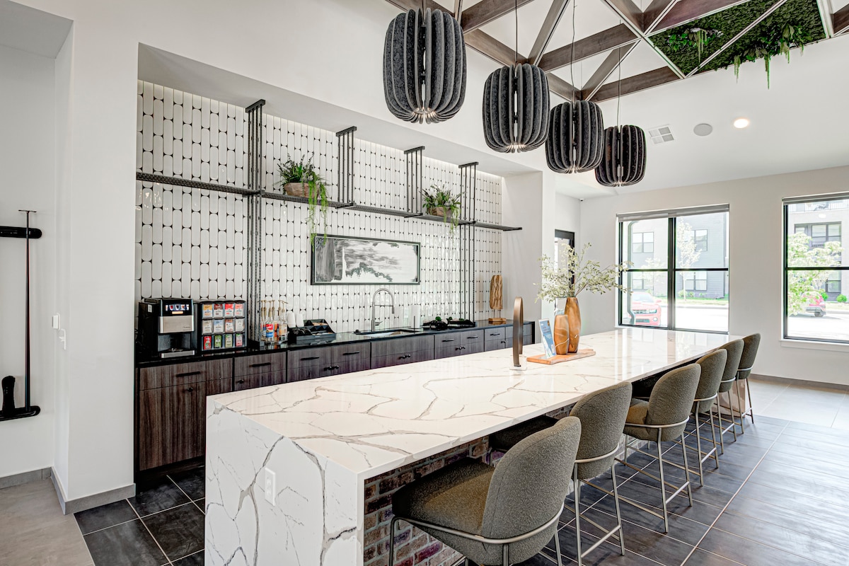 A community clubhouse kitchen area is presented with a long, white marble countertop and dark cabinetry. Modern pendant lights hang from the ceiling, illuminating the space. Plants are integrated into the design, adding a touch of greenery. A large window allows natural light to fill the room.