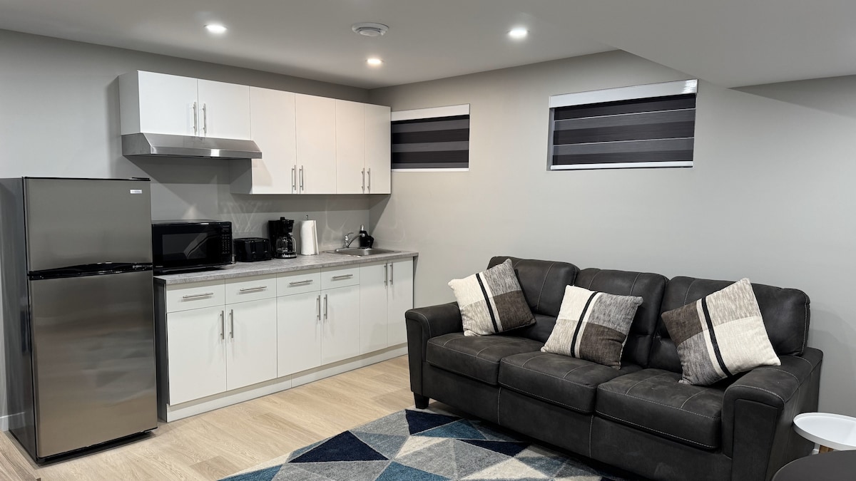 The living area presents a modern kitchenette equipped with a refrigerator, microwave, and coffee maker. A dark gray couch adorned with decorative pillows is positioned on a patterned area rug, while sleek cabinets line the wall above the countertop. Light enters through stylishly covered windows.