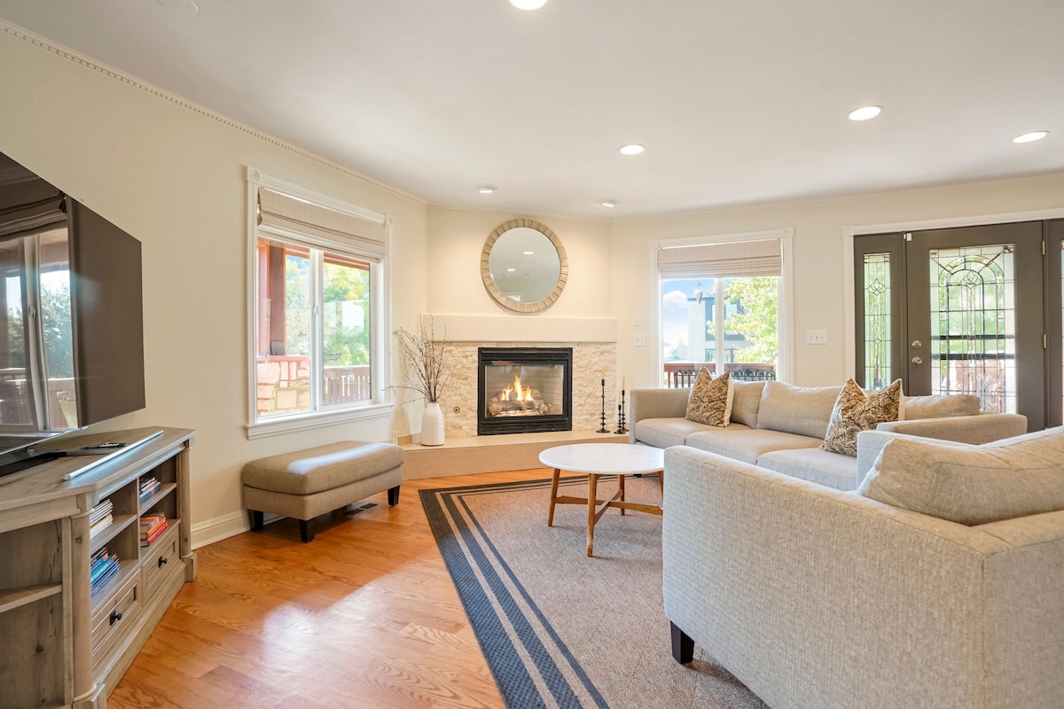 A cozy living area features a modern fireplace framed by a neutral color palette. Soft seating arrangements are positioned around a circular coffee table, with a large television mounted on a console. Natural light fills the space through multiple windows, enhancing the inviting atmosphere.