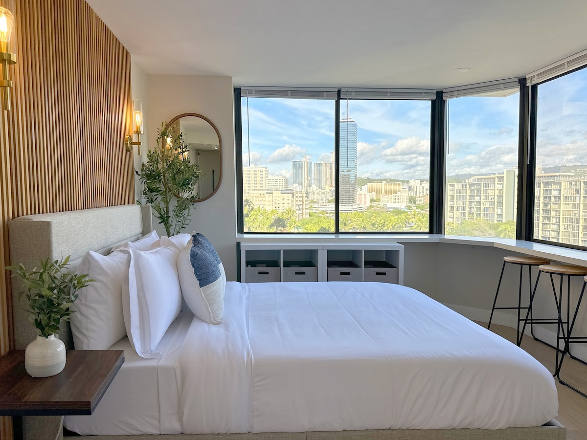 A bright and airy bedroom features a spacious bed with plush bedding. Large windows provide expansive views of the city skyline, allowing natural light to fill the room. A round mirror and decorative plants add subtle elegance to the contemporary design.