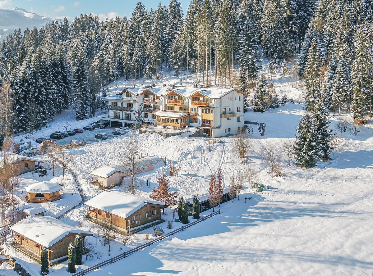 A surrounding snowy landscape encompasses a series of cozy wooden lodges and a multi-story building. The tranquil scene is enhanced by tall evergreen trees, creating a serene winter atmosphere. A clear blue sky complements the fresh snow covering the ground and rooftops.