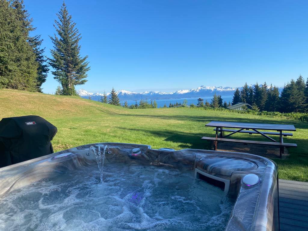 A hot tub is positioned on a private deck, surrounded by lush greenery. In the background, a panoramic view of Kachemak Bay and distant mountains is visible, under a clear blue sky. A large picnic table can also be seen on the deck.