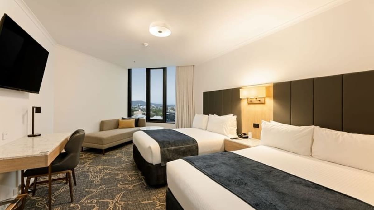 A spacious room features two large beds with dark blue throws, complemented by neutral-toned walls. A flat-screen TV is mounted on the wall, and a desk with a lamp is positioned beside a cozy seating area. Large windows invite natural light and offer views of the surroundings.