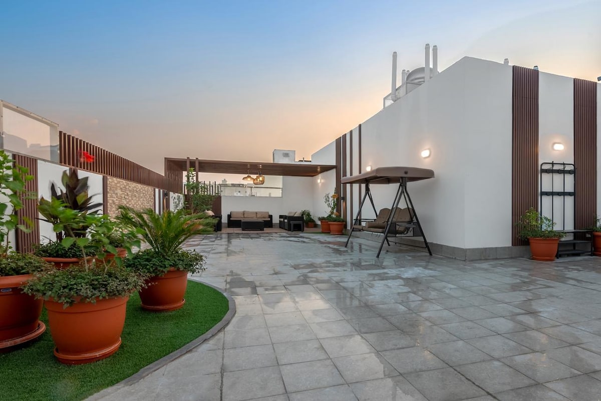 A spacious rooftop terrace features modern seating arrangements and a swinging chair, surrounded by potted plants. The surface is tiled, and the space is illuminated by soft lighting, creating a relaxed atmosphere as sunset hues fill the sky.