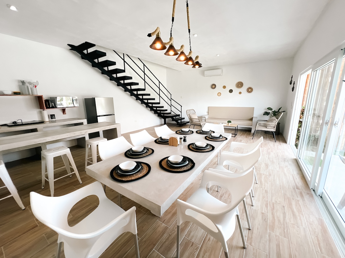 A spacious dining area is furnished with a large table surrounded by white chairs. A staircase with a metal railing leads to an upper level. Soft lighting fixtures hang from the ceiling, and large glass doors open to a patio, allowing natural light to fill the space.