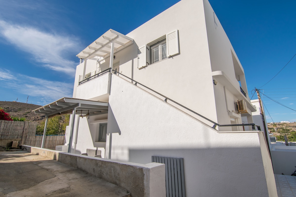 A modern two-story residence is depicted, featuring a white exterior with contrasting grey accents. The building is framed by a clear blue sky and showcases an outdoor staircase leading to an upper terrace, which is partially shaded by a white awning.
