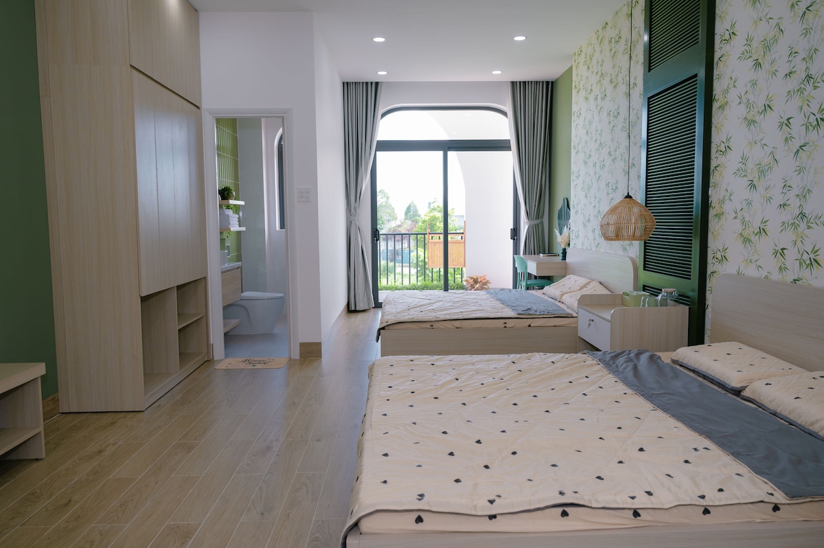 A modern sleeping area features two spacious beds with neutral-toned bedding. Natural light filters through large glass doors leading to a balcony, while decorative wallpaper adds a fresh touch. Minimalist furniture, including a wardrobe and nightstands, contributes to the room's serene atmosphere.
