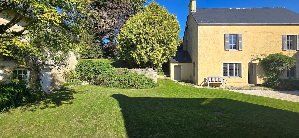 A well-maintained garden surrounds a historic yellow farmhouse, accented by green shrubs and trees. A wooden bench sits on the grassy area, with the sun creating distinct shadows across the lawn. The clear blue sky adds to the tranquil ambiance of the setting.