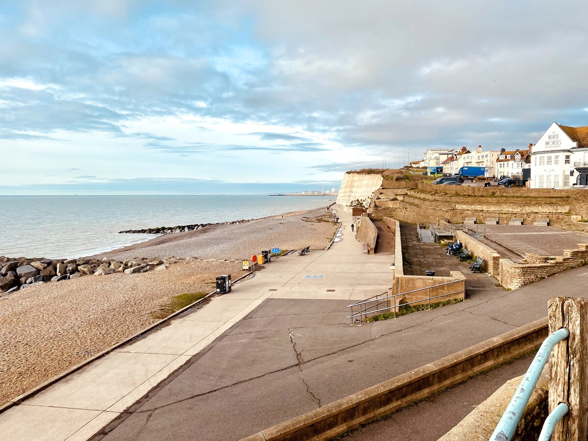 The image captures a calm beach scene with a wide stretch of pebbled shore and a gently lapping sea. A concrete walkway leads towards the water's edge, bordered by rocky formations. Buildings dot the background, maintaining a low profile amid the coastal landscape.