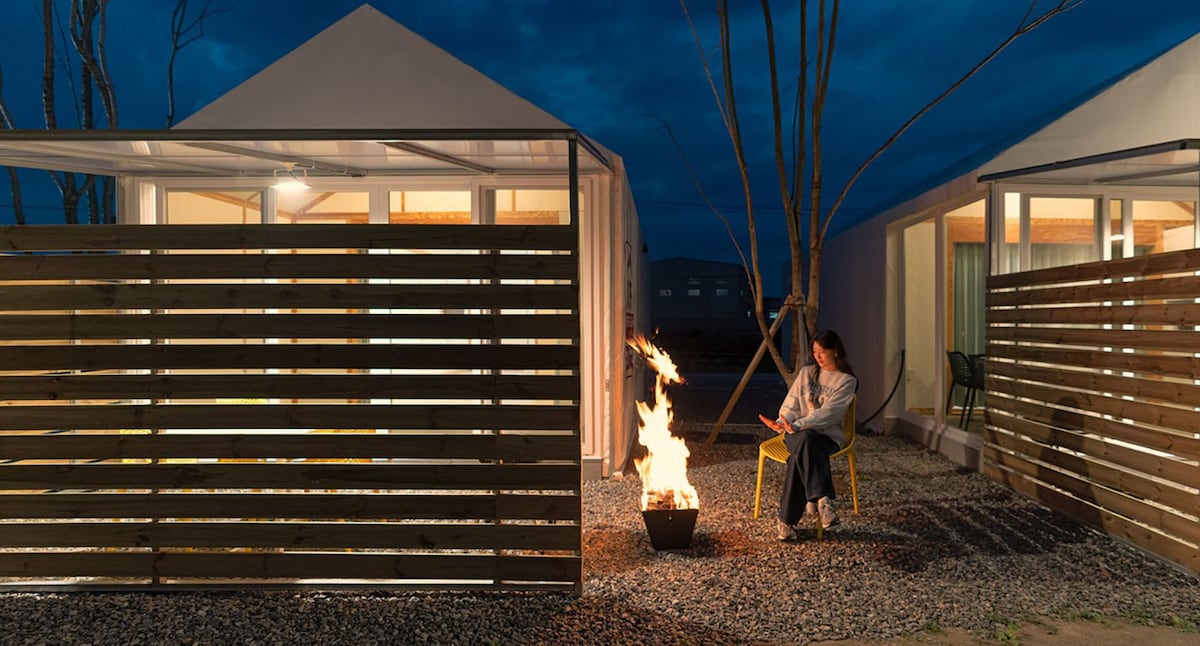 A serene evening setting features a cozy fire pit surrounded by a gravel area, with wooden fences providing privacy. Two cabins with large windows are visible in the background, softly illuminated by indoor lights, enhancing the calm atmosphere.