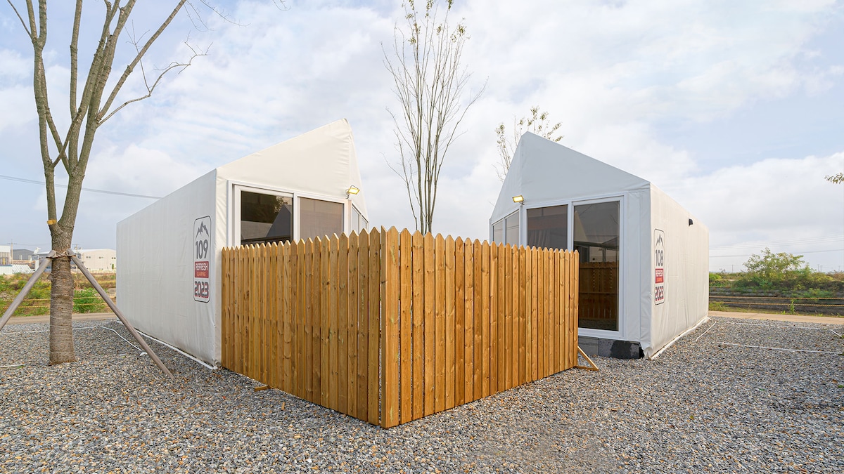 Two white glamping tents stand side by side, enclosed by a wooden fence. The gravel surface surrounds the tents, and the area appears spacious with trees in the background and a cloudy sky above.