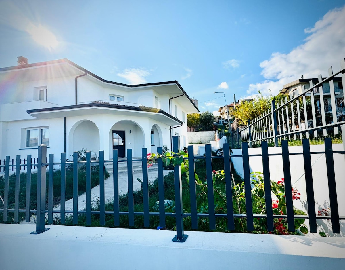 A modern villa is viewed from the exterior, featuring a white façade and arched entrances. A decorative iron fence surrounds the property, which includes landscaped areas with greenery and flowers. The sky is clear, contributing to a bright and inviting atmosphere.