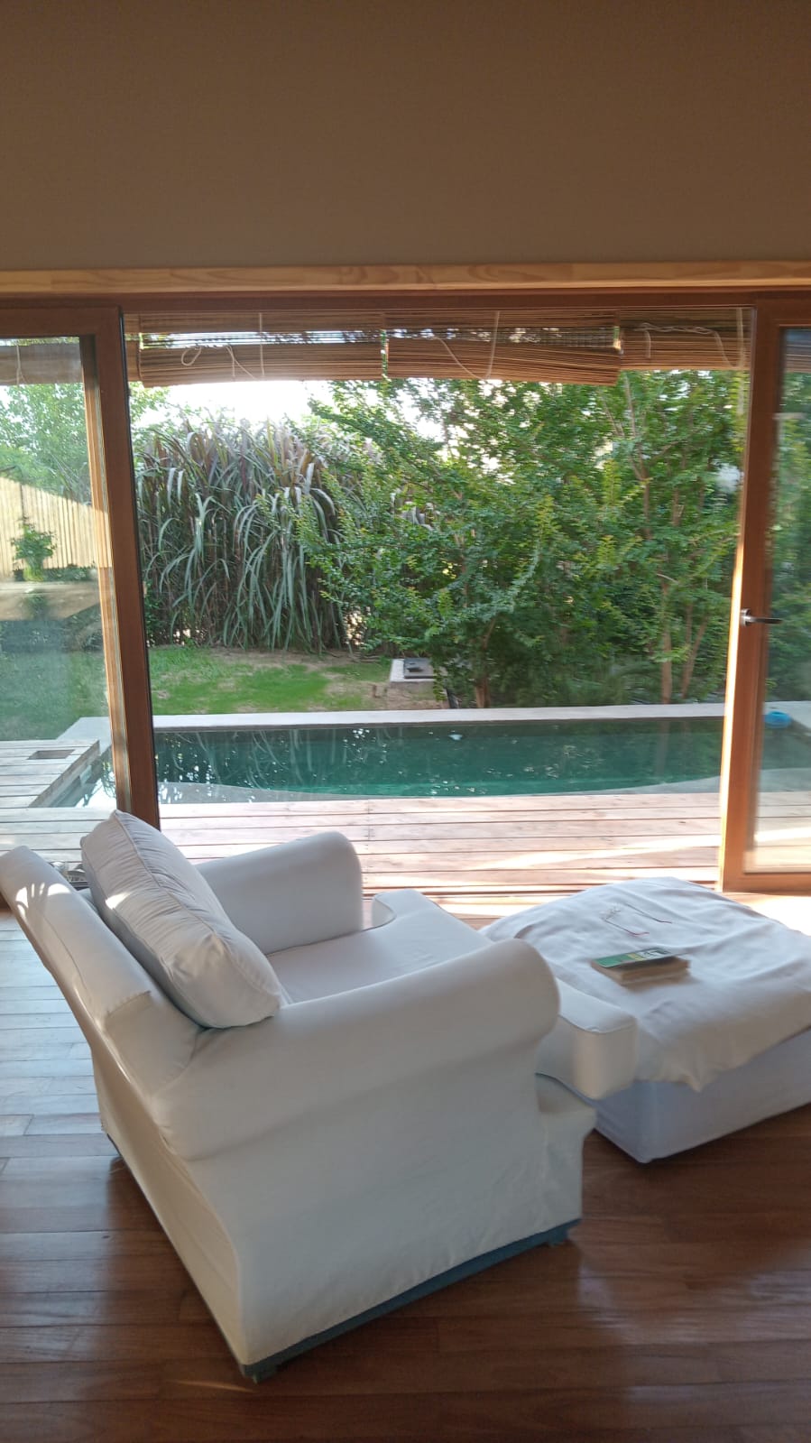 A bright seating area is framed by large glass doors that open to a serene outdoor view. A plush white chair sits alongside a matching ottoman, while sunlight illuminates the wooden deck and the inviting pool outside, surrounded by lush greenery.