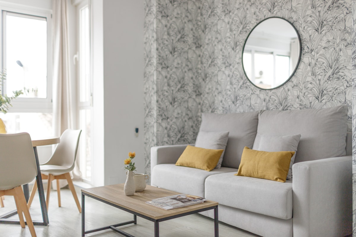 A light-filled living area features a comfortable gray sofa adorned with yellow cushions, placed in front of a circular mirror. A wooden coffee table holds a small flower vase, while a dining table with light-colored chairs is visible in the background.