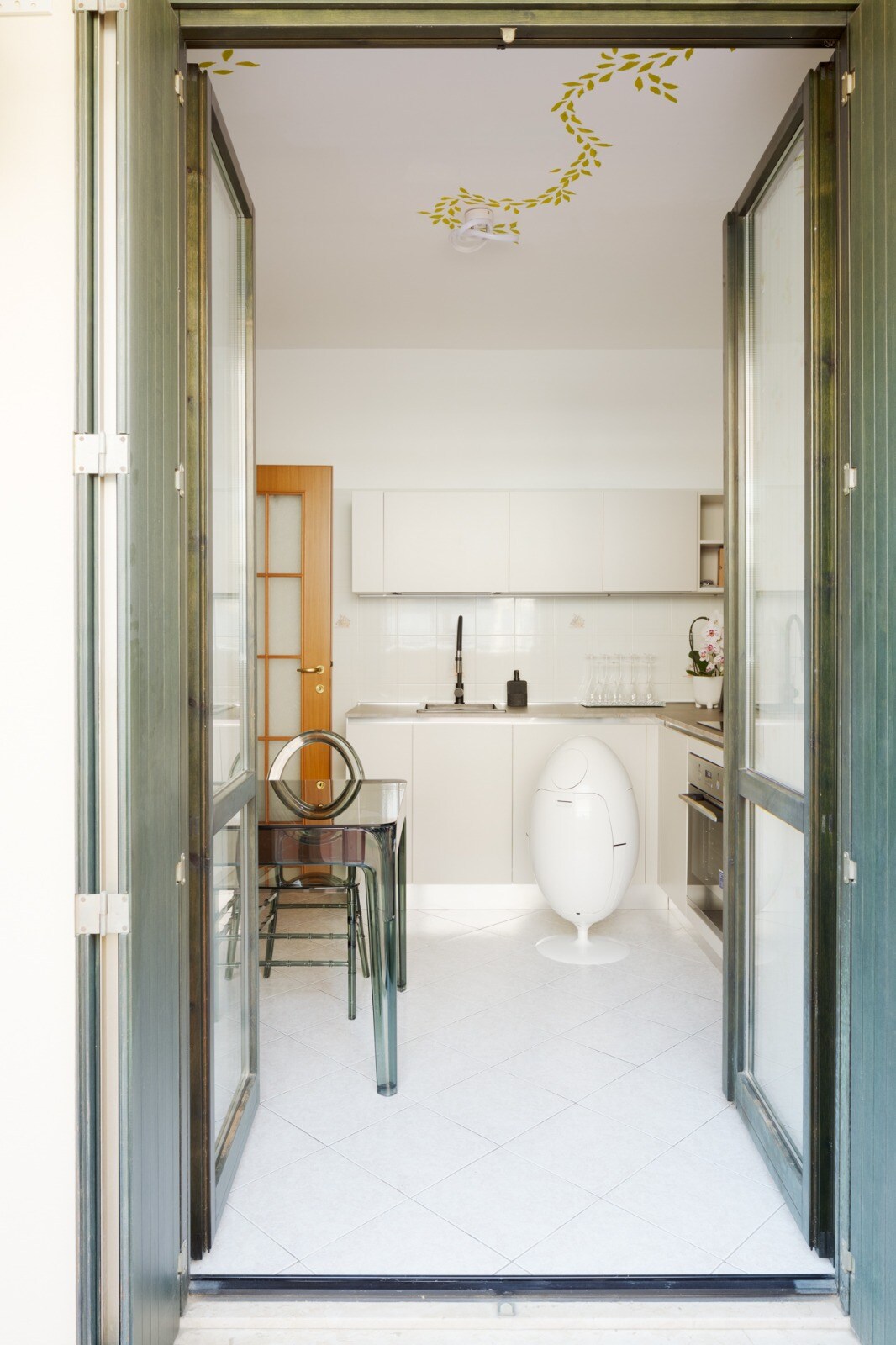 A bright kitchen area is visible through open glass doors. The space features modern cabinetry, a sleek countertop, and a dining table with chairs. Natural light illuminates the tiled floor, creating a welcoming environment. Decorative elements add a touch of charm to the minimalistic design.