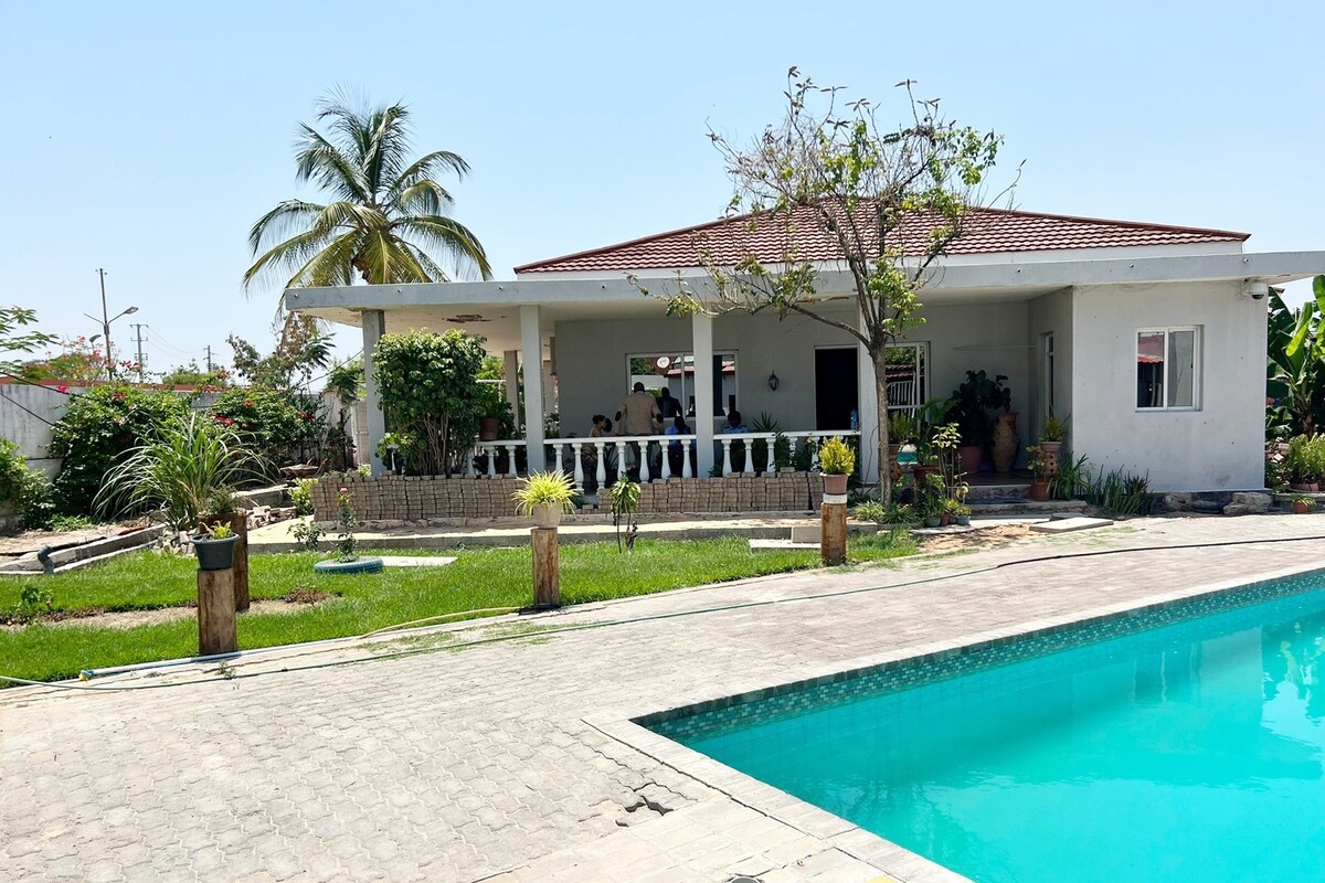 A serene outdoor area features a clear swimming pool reflecting the sky. Adjacent, a single-story building showcases a spacious porch with multiple columns, surrounded by well-maintained gardens and tropical plants. The scene is set against a bright and clear backdrop.