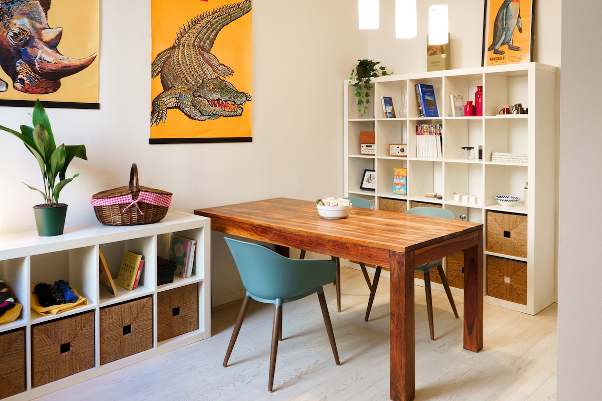 A dining area features a wooden table accompanied by two contemporary chairs. Colorful animal artwork adorns the walls, while a bookshelf displays various items and decorative accents. A potted plant adds a natural touch to the space, creating a welcoming atmosphere.