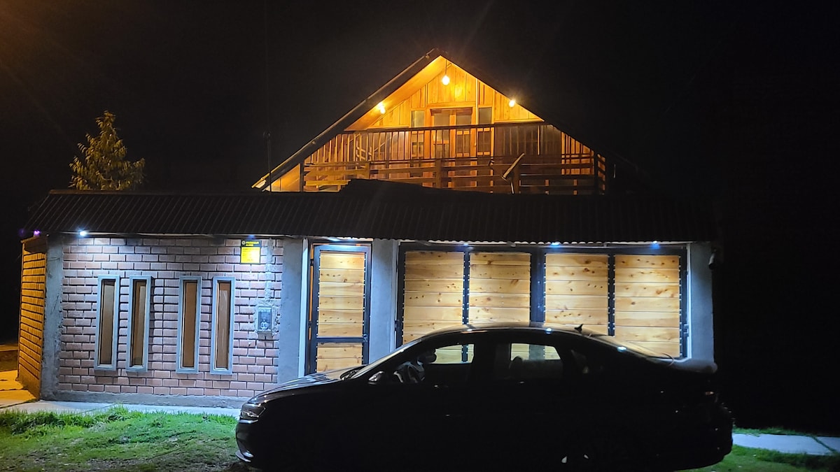 The exterior of the house is illuminated at night, showcasing a combination of wooden beams and stone accents. A spacious balcony extends above the entrance, and a parked car is visible in the foreground, framing the inviting atmosphere of the property.