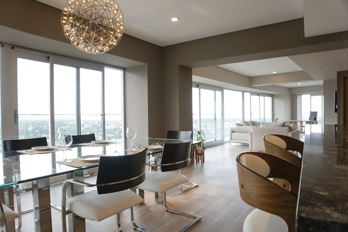 A modern dining area features a glass tabletop surrounded by six sleek chairs. Large windows line the wall, providing expansive views of the cityscape. A stylish light fixture hangs from the ceiling, illuminating the space with a soft glow, while a comfortable seating area is visible in the background.