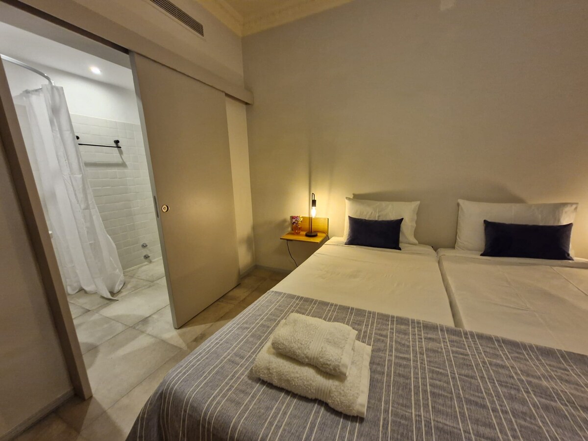 The room features two spacious beds neatly arranged, complemented by decorative cushions. A nightstand with a lamp is positioned beside one bed. A partially open door leads to a bathroom area, which includes a shower with a curtain and tiled flooring.