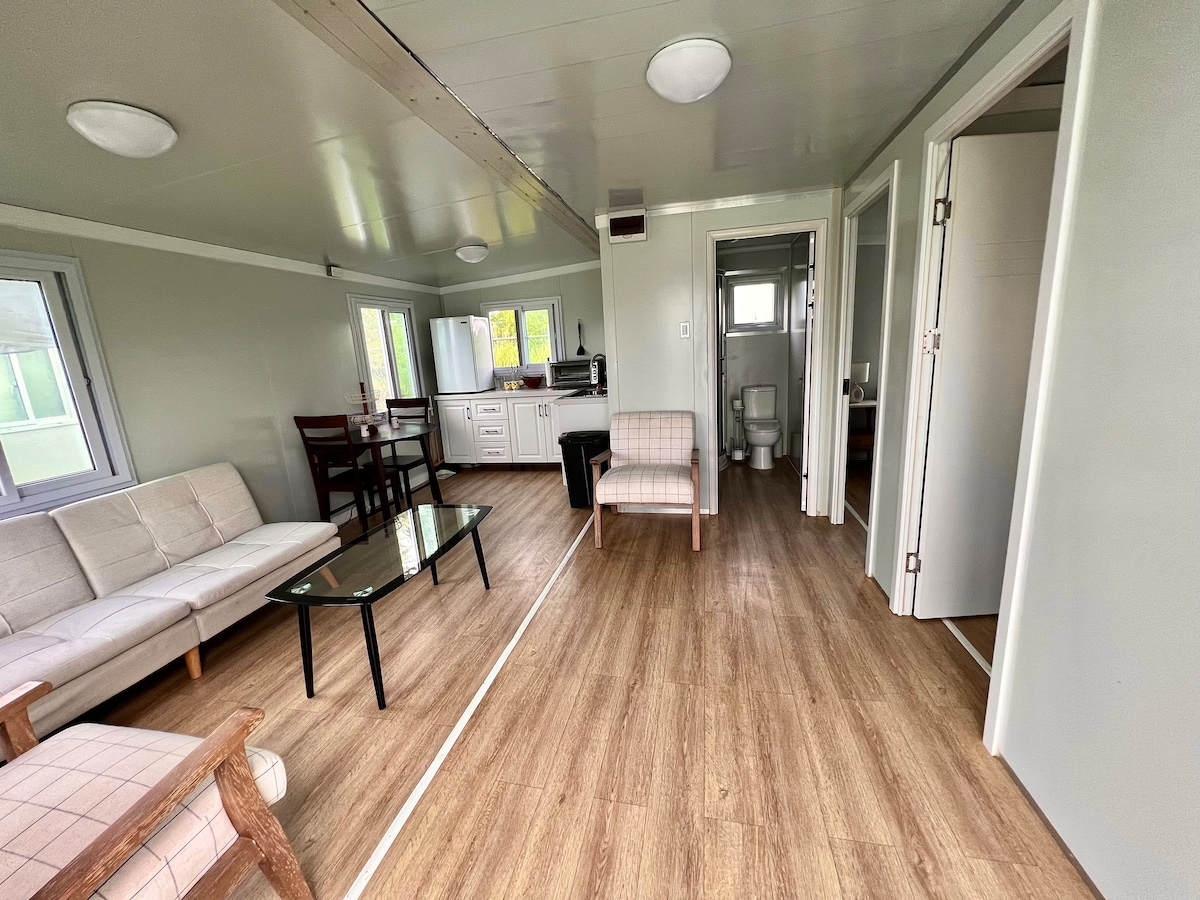 An open living area is shown, featuring a light-colored sofa and two chairs arranged around a glass coffee table. A kitchenette is visible in the background, equipped with a sink and counter space. Natural light enters through multiple windows, illuminating the wooden flooring.