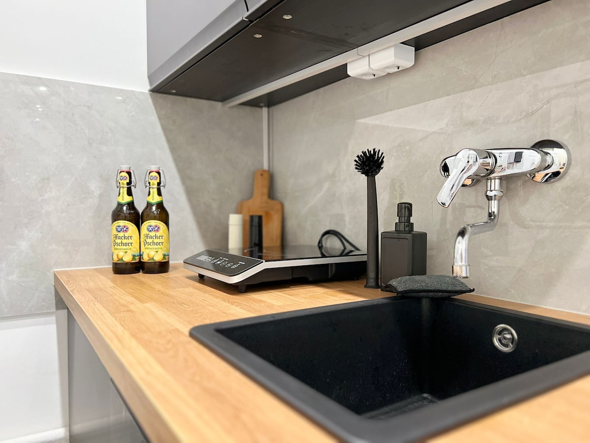 A modern kitchen area features a clean wooden countertop with a black sink. A soap dispenser and cleaning brush are positioned near the sink. Two bottles are placed next to a wooden cutting board, with a sleek induction cooktop incorporated into the space.