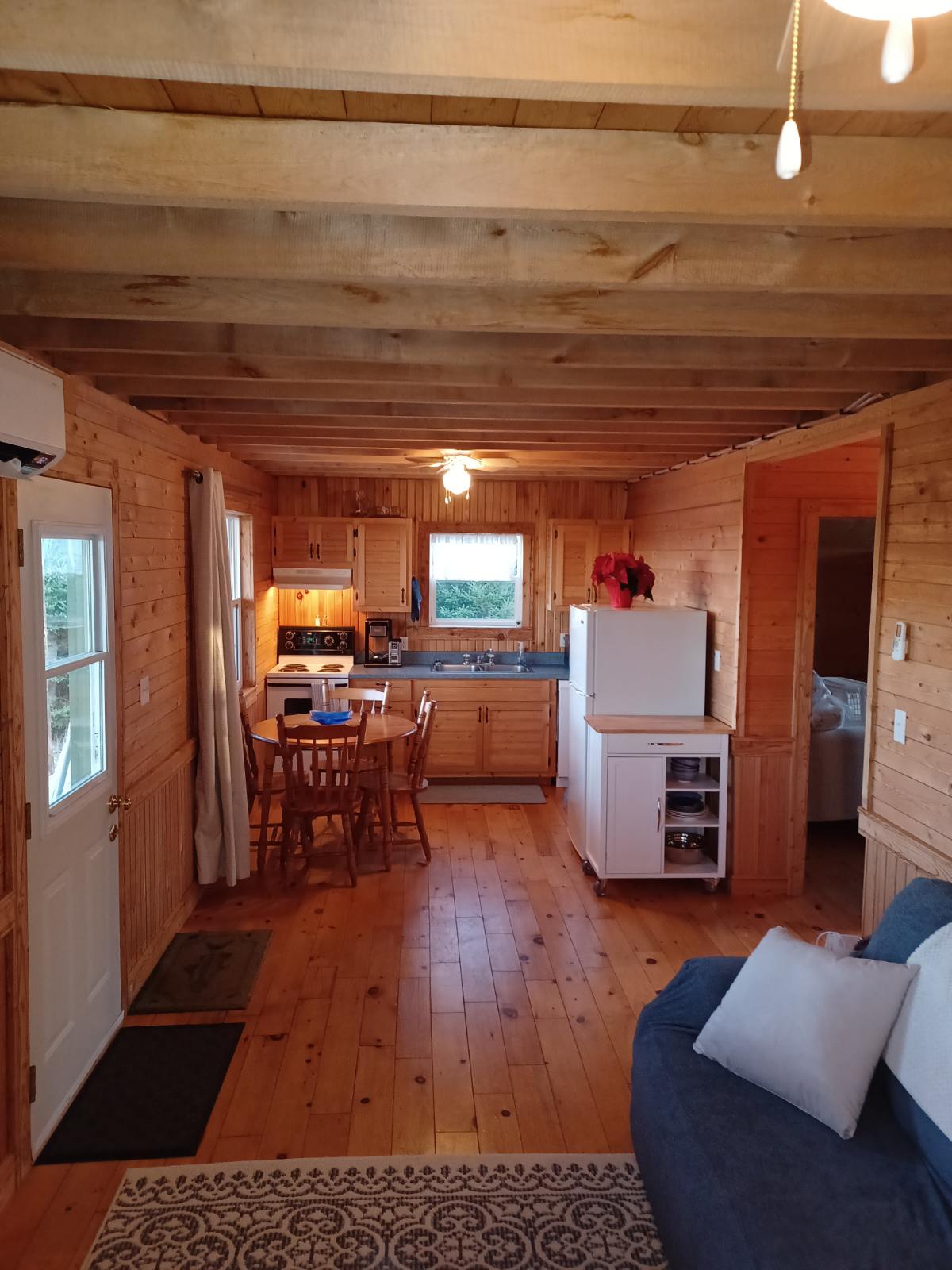 A cozy interior featuring wooden walls and ceiling beams. The kitchen area includes an oven, stove, and refrigerator, with a dining table surrounded by four chairs. A comfortable couch with cushions is visible, complemented by a decorative area rug on the warm wooden floor.