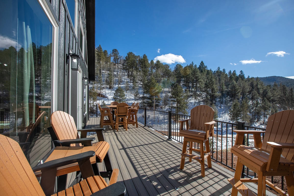 A private deck features wooden seating arranged for group gatherings, overlooking a snowy landscape filled with trees. Sunlight illuminates the scene, offering a serene view of the surrounding mountains and nature.