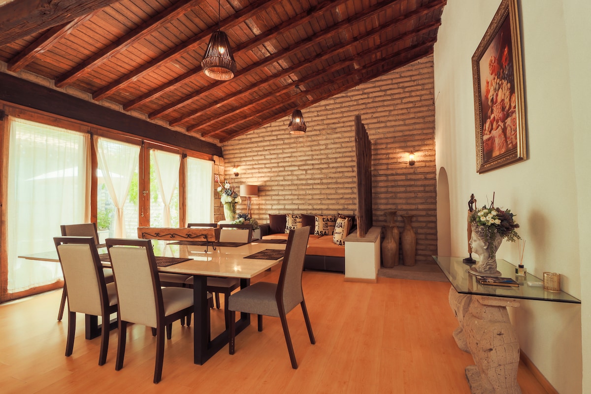 An inviting living area showcases high wooden beams and a warm, natural color palette. A large dining table is surrounded by six chairs, while a comfortable seating arrangement is found in the background. Large windows allow ample natural light to fill the space.