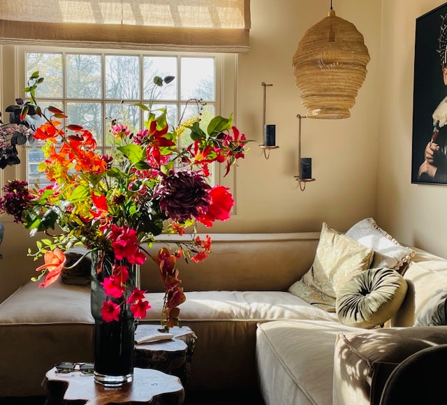 A well-lit living area features a plush corner sofa adorned with decorative pillows. A large vase filled with colorful flowers creates a vibrant focal point. Natural light streams through a large window, highlighting the textured wall hanging and artistic decor.