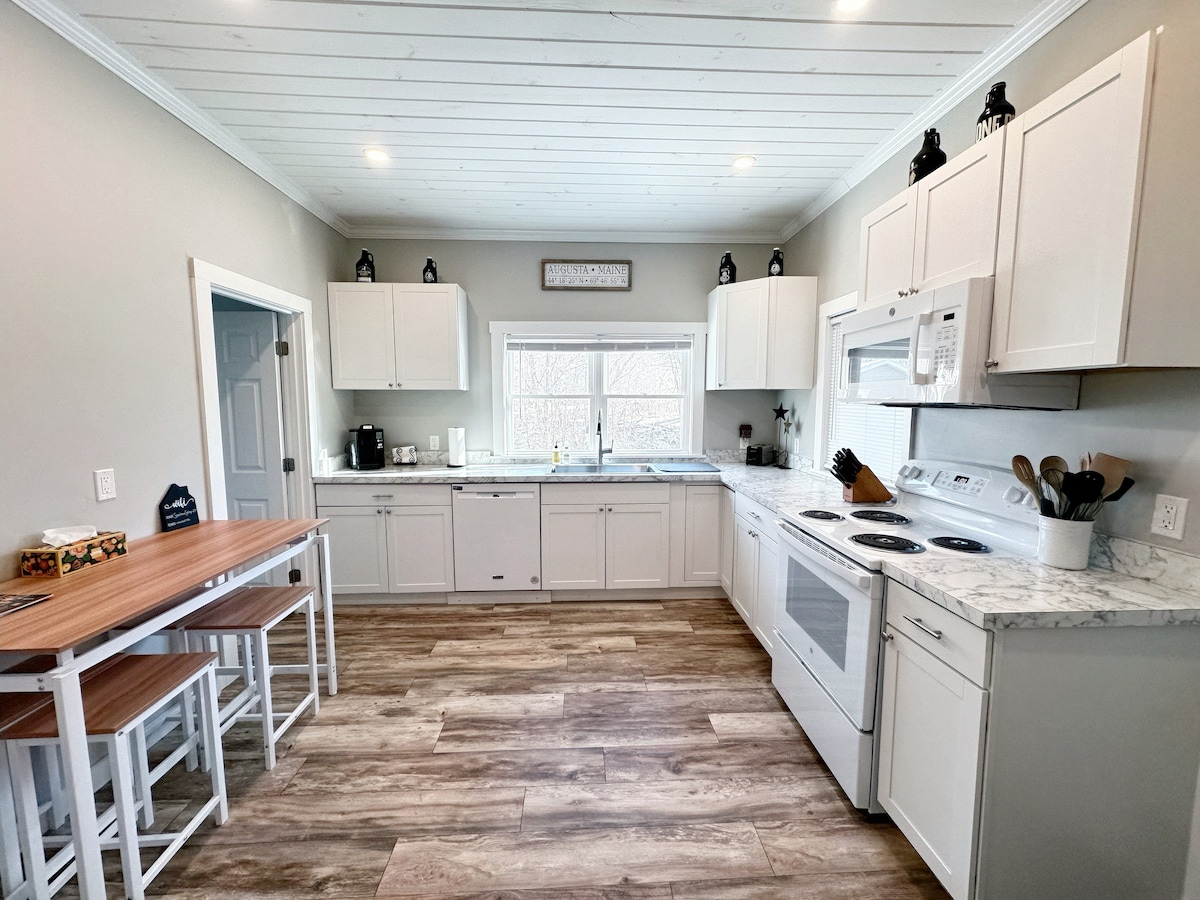 A modern kitchen features white cabinetry and countertops, complemented by wood and white bar stools at a small dining table. Appliances include an oven, microwave, and refrigerator. Natural light enters through a large window, illuminating the space and highlighting the seamless layout.