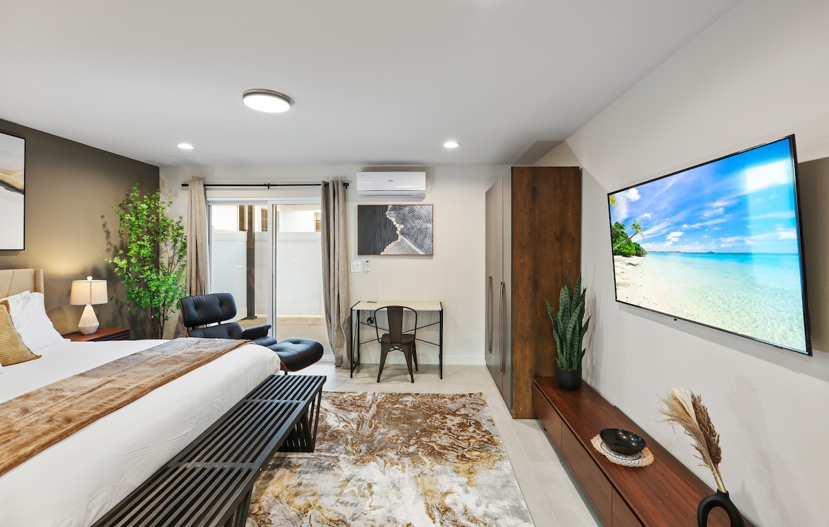 A spacious room features a king bed with neutral bedding and a decorative throw. A large flat-screen TV is mounted on the wall opposite a sleek desk and chair. Natural light floods in from sliding glass doors, which provide access to a private patio.