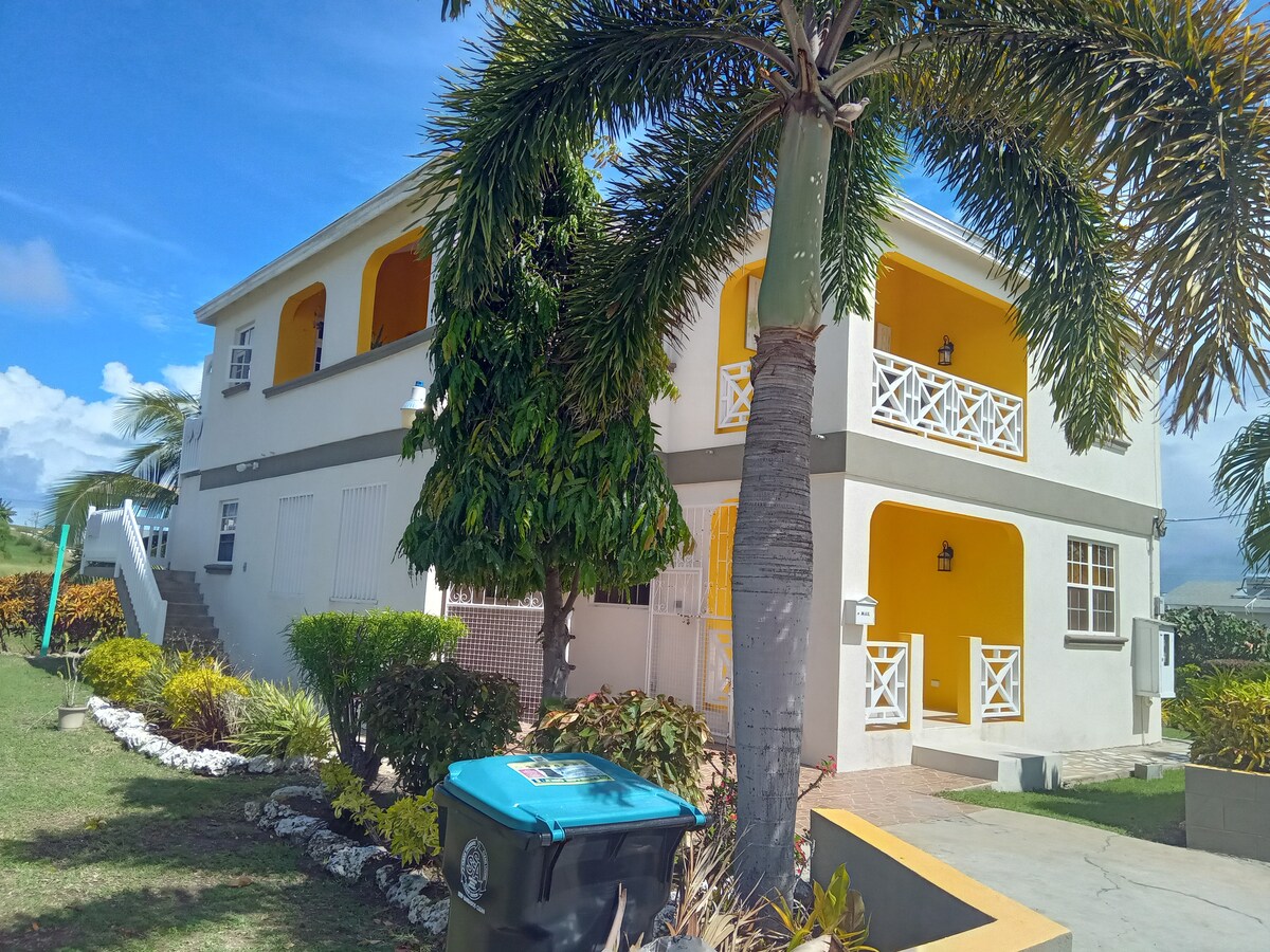 A charming two-story building with bright yellow accents is surrounded by lush greenery and colorful landscaping. The exterior features white railings and a series of steps leading up to the entrance. A large palm tree adds a tropical touch to the inviting atmosphere.