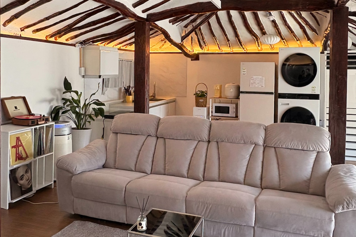 A spacious living area features a comfortable sofa under wooden beams. A subtle warm light runs along the ceiling, enhancing the cozy ambiance. Nearby, modern appliances including a washing machine and microwave are visible, complemented by a small plant and decorative items.