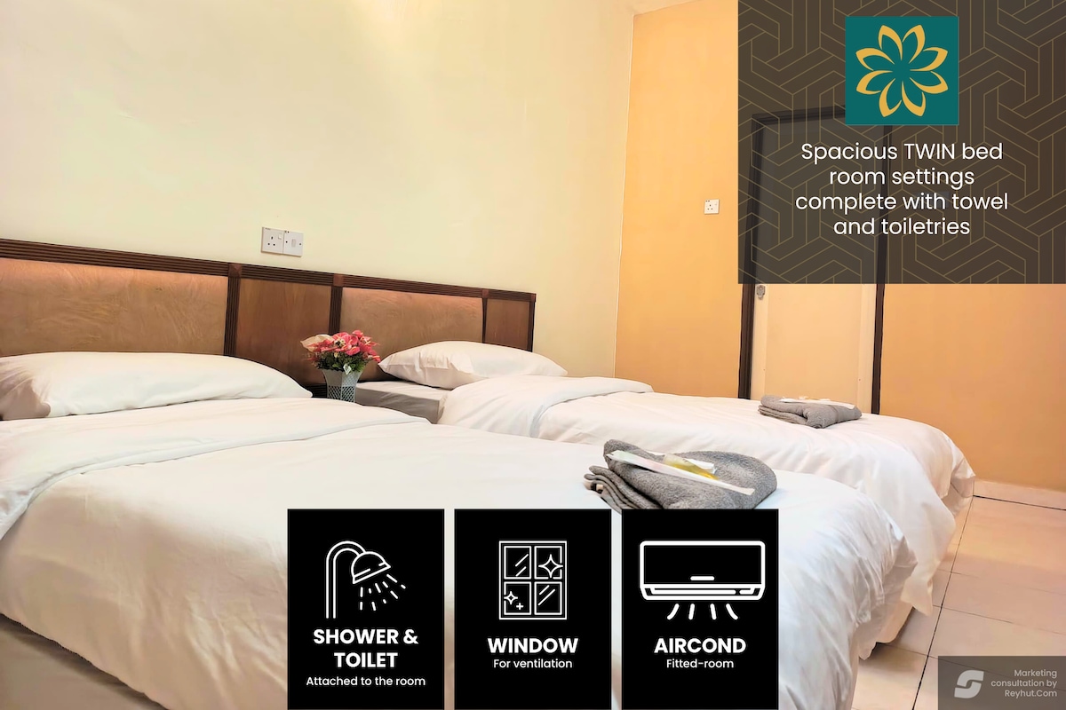 A twin bedroom features two spacious beds with neutral bedding. A small bouquet sits on a bedside table. A window provides natural light, and an air conditioning unit is visible. The room includes an attached shower and toilet, with towels and toiletries neatly arranged.