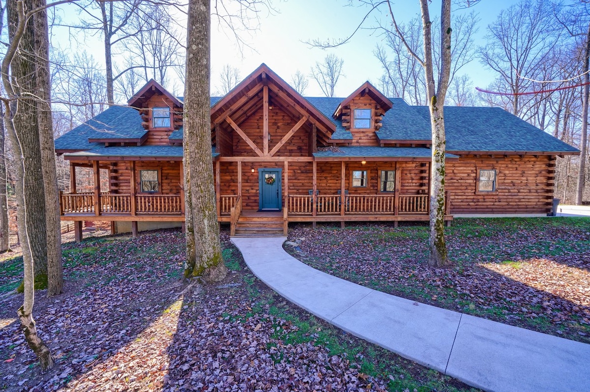 A custom-built log cabin is surrounded by tall trees and natural foliage. The entrance features a welcoming blue door, framed by a spacious porch with wooden railings. A concrete path leads to the entryway, enhancing accessibility to the home.