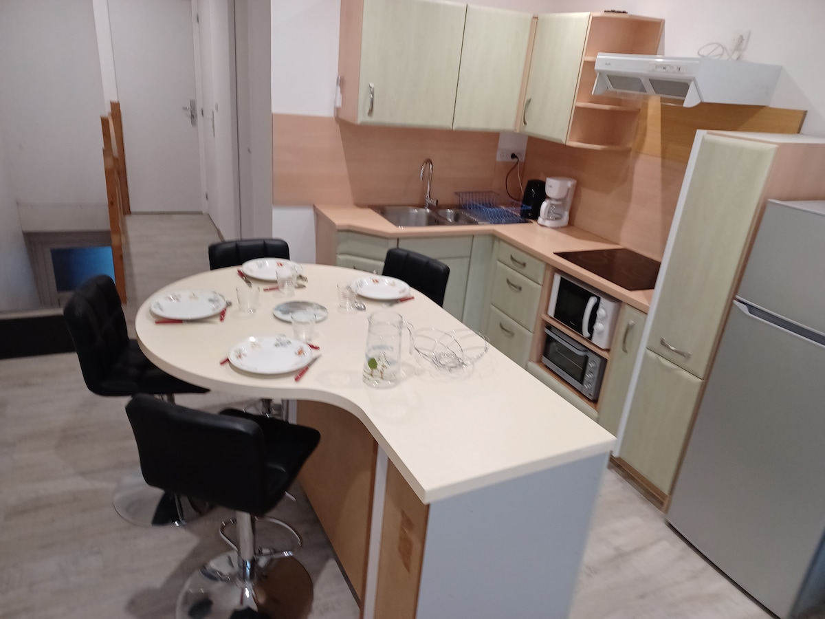 A modern kitchen features light-coloured cabinetry and a curved breakfast bar with four black stools. A stovetop and microwave are present along with a refrigerator and coffee maker, creating a functional cooking space. Plates are neatly arranged on the counter, ready for use.