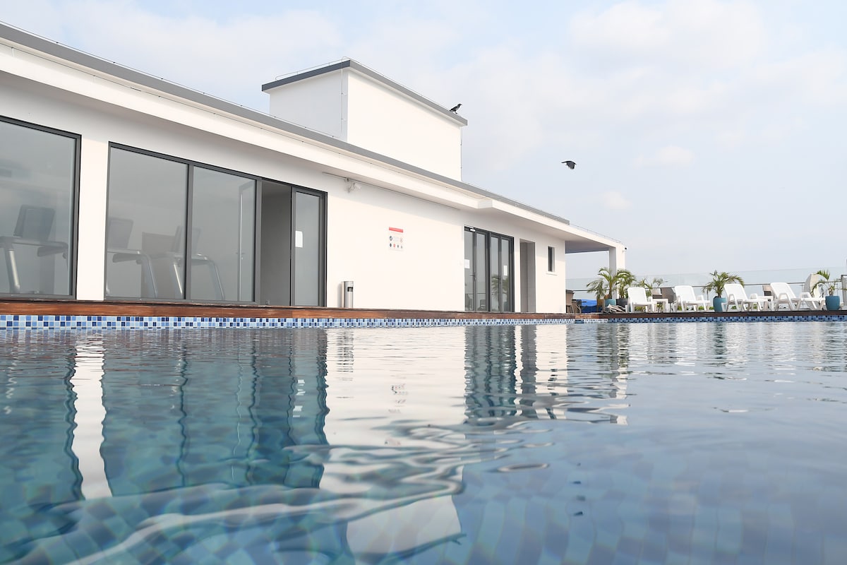 A serene swimming pool is visible, reflecting the blue sky above. The pool is bordered by a wooden deck, with modern glass doors leading to a fitness area. Comfortable lounge chairs are arranged around the pool, inviting relaxation amid the peaceful setting.