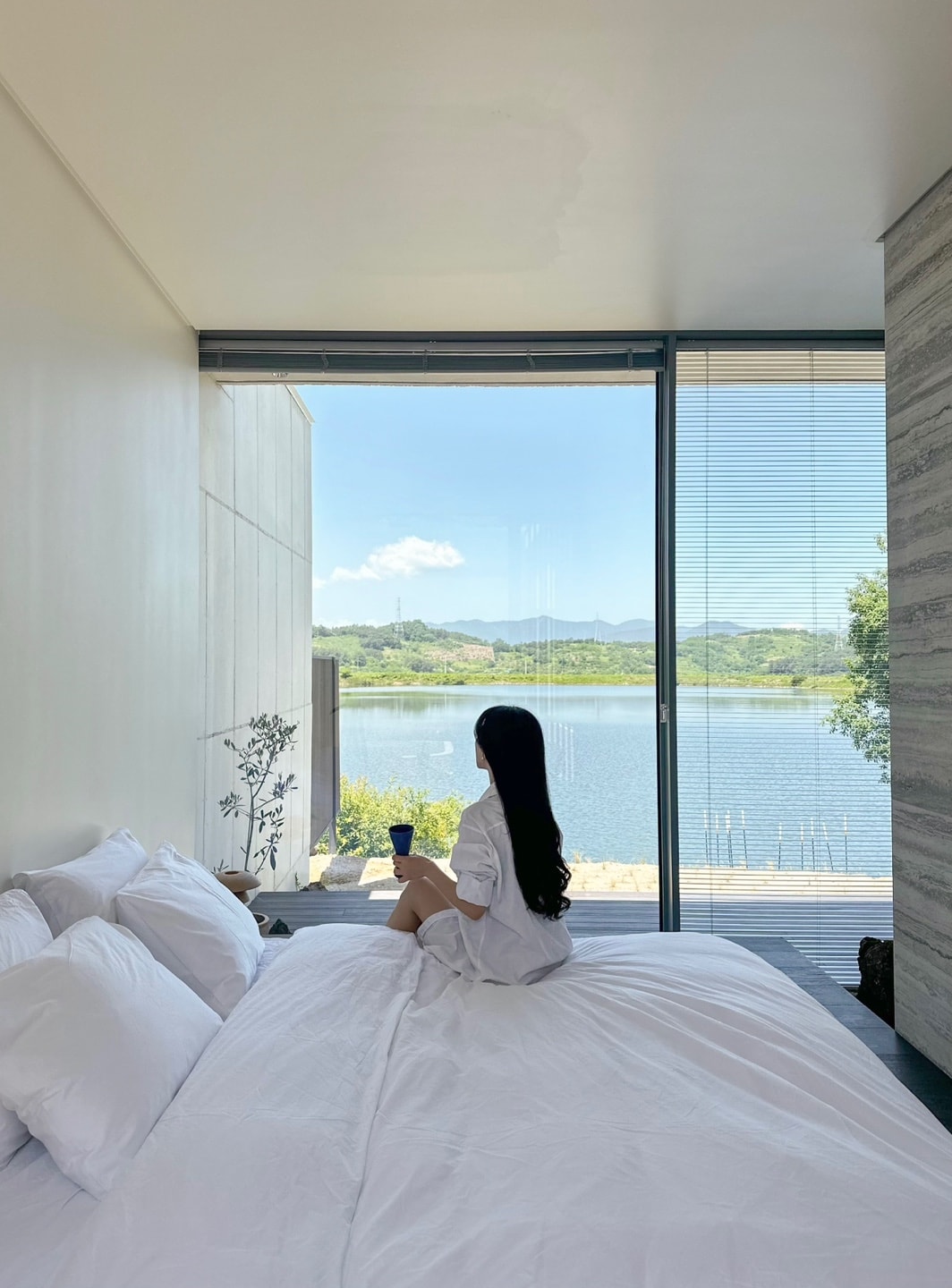 A bright bedroom features a spacious bed with white linens, positioned near large glass windows overlooking a serene lake and lush greenery. Soft natural light fills the room, enhancing the view of outdoor landscapes and distant mountains.