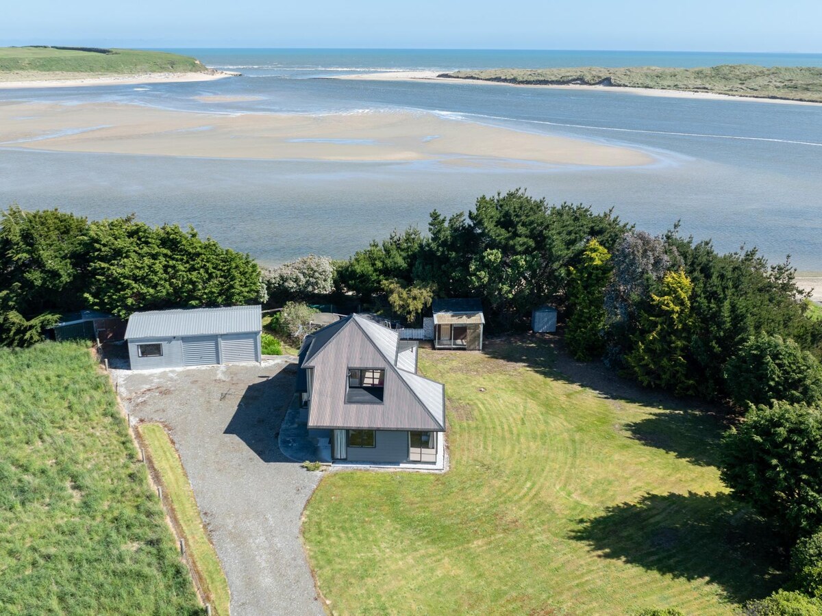 An aerial view of a spacious property near an estuary, surrounded by greenery. The house features a modern roof and large windows, while the landscape includes open grass areas and clusters of trees. The tranquil water and sandy beach are visible in the background.