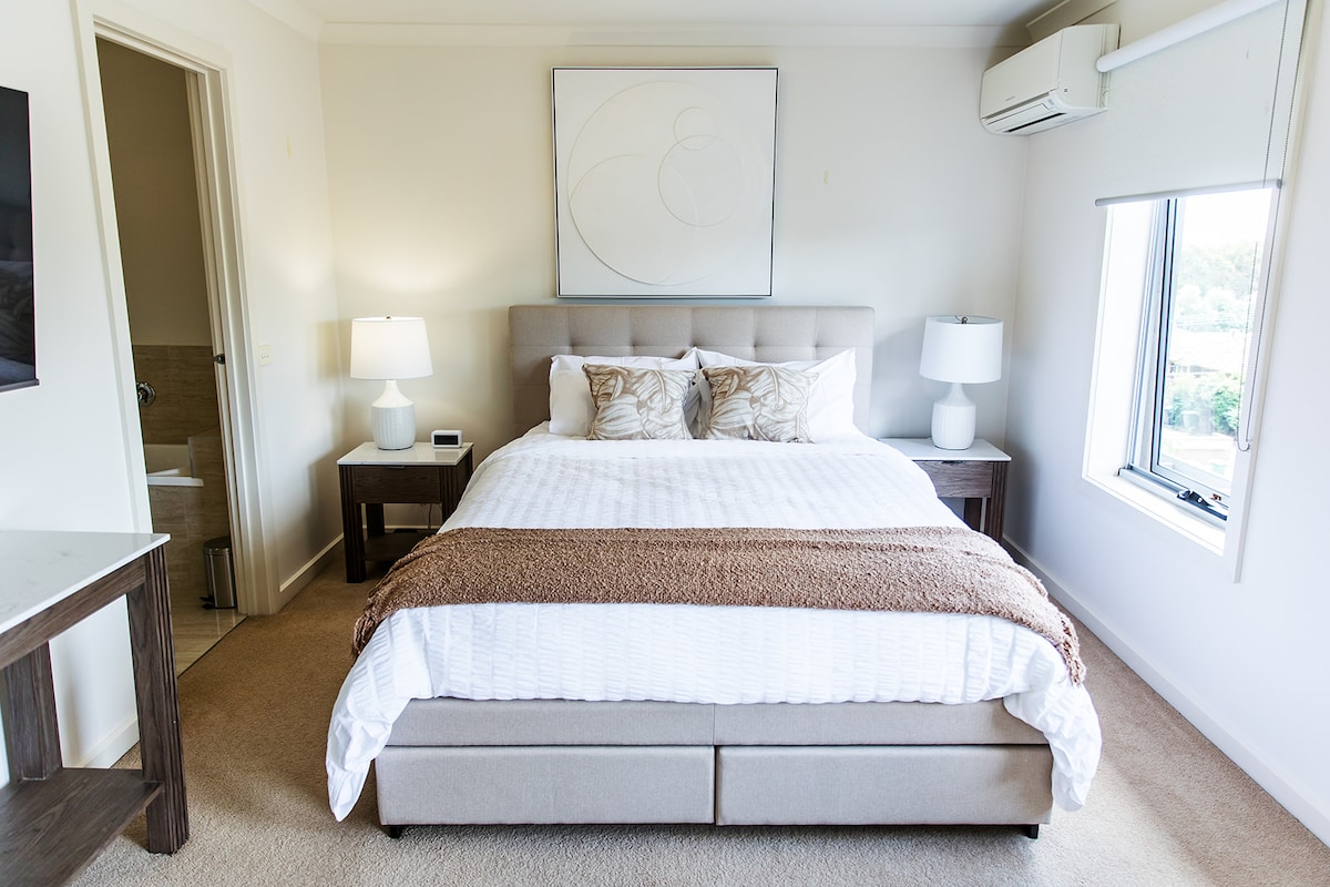 A well-appointed bedroom features a comfortable queen-size bed with neutral linens and decorative pillows. Soft lighting is provided by two bedside lamps on dark wood nightstands. A large window allows natural light to fill the space, while a modern artwork hangs above the bed.