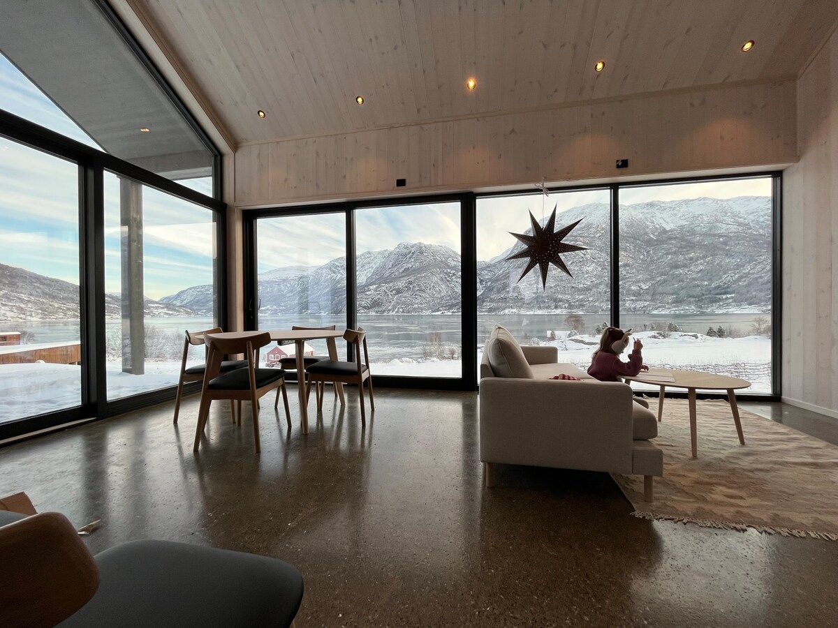A spacious living area is highlighted by large panoramic windows offering views of snow-covered mountains and a serene fjord. Modern furniture includes a light-colored sofa and a dining table. A decorative star hangs from the ceiling, adding a touch of warmth to the bright interior.